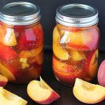 two jars filled with pickled fruit sitting on top of a wooden table next to sliced peaches