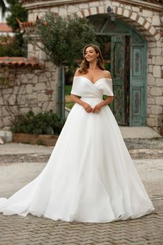 a woman in a white wedding dress standing on a cobblestone street with her hands behind her back