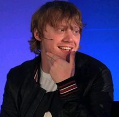 a young man smiles while sitting in front of a blue screen with his hand on his chin