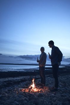 two men standing next to a campfire on the beach at dusk with water in the background