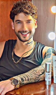 a man with tattoos sitting at a table in front of a mirror and bottle of deodorant