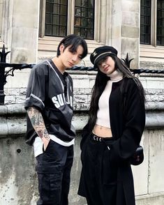 two young people standing next to each other in front of a stone wall and building