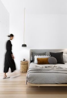 a woman walking past a bed in a room with white walls and wooden floors,