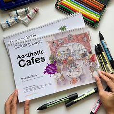 a person holding a coloring book over a table with markers and crayons on it