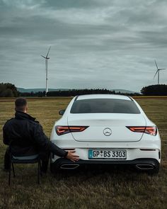 a man sitting in a chair next to a white car with windmills in the background