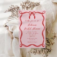 a pink wedding card with a red ribbon on it next to some dried flowers and white fabric