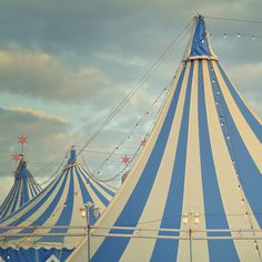 a large blue and white circus tent with lights on it's sides, in front of cloudy sky