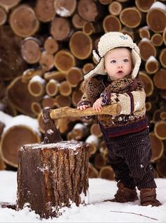 a small child standing next to a pile of logs in the snow holding a wooden stick