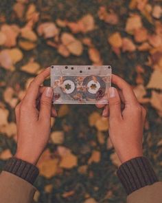 someone holding up a cassette in front of some leaves and flowers on the ground with their hands