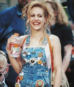 a woman in overalls holding a bottle and smiling