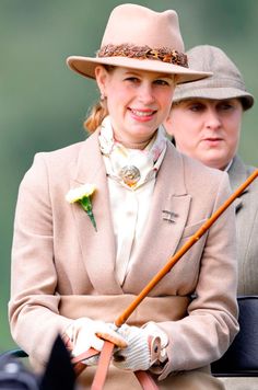 a woman in a tan coat and hat holding a brown stick while standing next to another woman