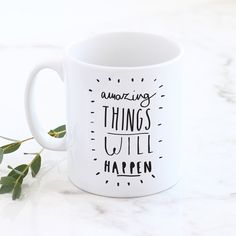 a white coffee mug with the words amazing things will happen written on it next to some green leaves