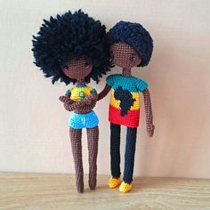 two crocheted dolls standing next to each other on a wooden table with a white wall in the background