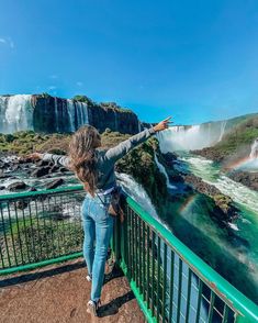 a woman standing on top of a metal railing next to a waterfall and rainbow in the sky