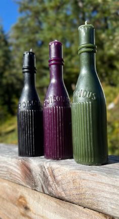 three different colored bottles sitting on top of a wooden table next to some pine trees