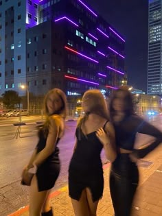 three beautiful young women standing next to each other in front of tall buildings at night