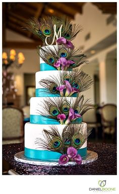 a wedding cake decorated with peacock feathers and purple flowers is displayed in front of a chandelier