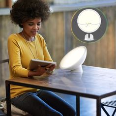 a woman sitting at a table reading a book with an electronic light above her head