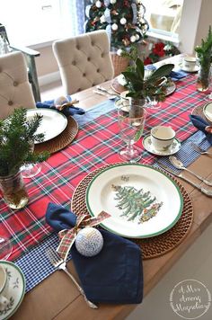 the table is set for christmas dinner with plates and place settings on it, along with evergreens