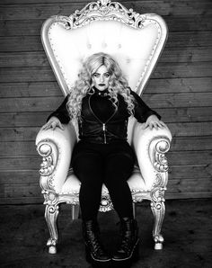 a woman sitting on top of a white chair in front of a wooden wall and wearing black