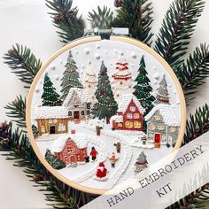 a cross stitch christmas scene with houses and trees in the snow, surrounded by pine needles