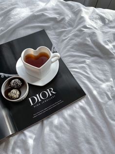 a book about dior is sitting on a bed next to a cup of tea