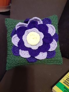 a crocheted pillow with a purple flower on it sitting on a table next to a book