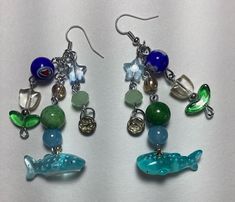 ❀ Handmade and one of a kind ❀ flower and sealife themed blue, green and silver wire wire earrings ❀ 18 round, star, and tulip  shaped beads ❀Great gift for friend, family member, or anyone who enjoys a little bit of joy and whimsy ❀ Dimensions: 3 1/2" length 2" wide (both earrings set side by side) Whimsical Blue Nickel-free Earrings, Whimsical Blue Charm Jewelry, Fun Blue Dangle Jewelry, Blue Fun Dangle Jewelry, Fun Blue Round Beads Jewelry, Whimsical Blue Jewelry With Charms, Blue Dangle Jewelry With A Fun Style, Fun Blue Round Beaded Jewelry, Handmade Blue-green Jewelry Gift