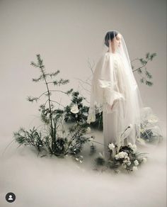 a doll dressed in white standing next to flowers and greenery on a gray background