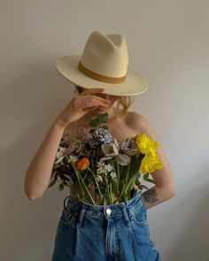 a woman wearing blue jeans and a white hat holds flowers in front of her face