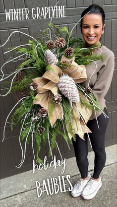 a woman standing in front of a garage door holding a wreath with pine cones and evergreens