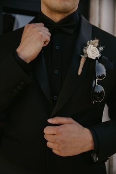 a man in a tuxedo adjusts his bow tie with sunglasses and a boutonniere