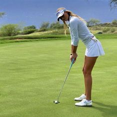 a woman in white shirt and skirt playing golf on green field with blue sky behind her