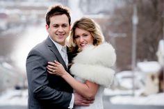 a man and woman hugging each other in front of snow covered street with buildings behind them