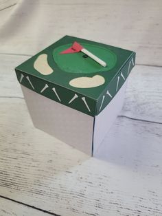 a green box with a red clock on the top sitting on a white wooden table