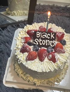 a white cake with strawberries and blueberries on it that says black stones next to a lit candle