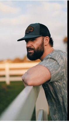 a man with a beard wearing a hat leaning on a rail in front of a white fence