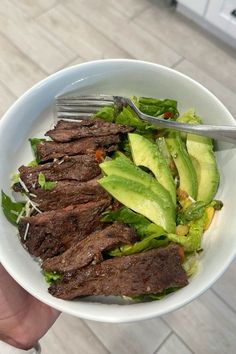 a person holding a white bowl filled with steak and lettuce topped with avocado