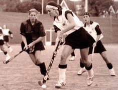 two women playing field hockey on a brick surface with other players in black and white