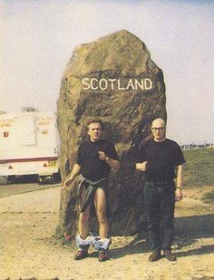 two men standing in front of a large rock with the word scotland on it and an rv behind them