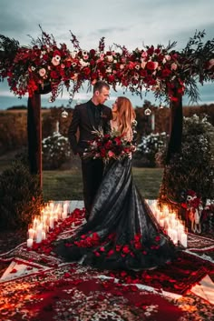 a man and woman standing under an arch with candles in front of them, surrounded by red flowers