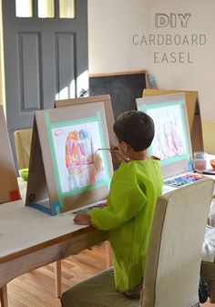 a little boy sitting at a table with some paintings on the easel and an easel in front of him