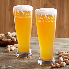 two beer glasses sitting on top of a wooden table next to peanuts and nuts in front of them