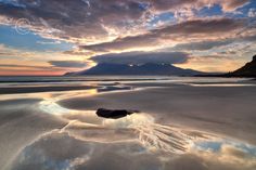 the sky is reflected in the wet sand on the beach at low tide, with mountains in the distance