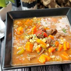 a bowl of soup with meat, carrots and other vegetables on a wooden table