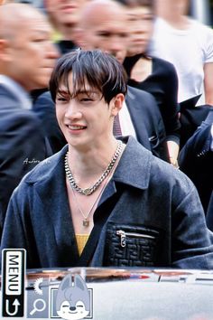 a young man wearing a black shirt and silver necklace standing in front of a group of people