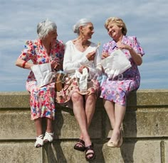 three older women sitting next to each other on top of a cement wall and talking