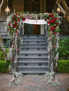 a wedding arch decorated with red flowers and greenery on the steps leading to an entrance