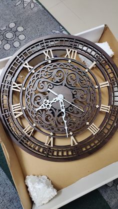 a large metal clock with roman numerals in a box on the floor next to a piece of paper