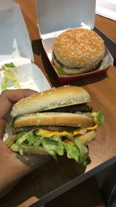 a person holding a hamburger in their left hand next to two takeout boxes on a table